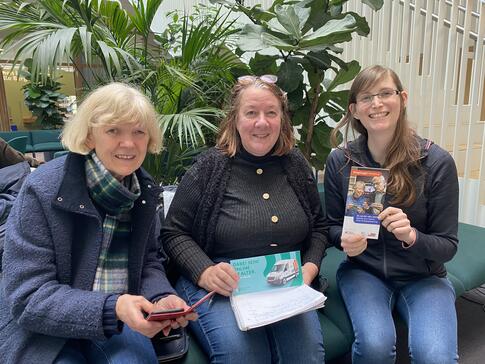 Petra (rechts) sitzt mit den beiden Freundinnen Astrid (Mitte) und Christine (links) im Atrium des K&ouml;rberHauses. Petra und Astrid halten zwei Flyer in den H&auml;nden und Christine ihr Smartphone.