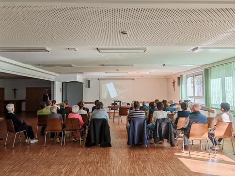 Zahlreiche Personen sitzen auf St&uuml;hlen im Raum verteilt und blicken nach Vorne zu einer Leinwand.
