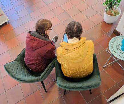 Petra (links) sitzt neben einer Seniorin mit einer gelben Jacke. Die beiden schauen sich gemeinsam ein Smartphone an. Das Bild ist aus der Vogelperspektive entstanden.