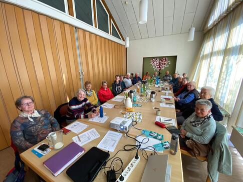 18 Senior:innen - davon die gro&szlig;e Mehrheit Frauen - sitzen an einem langen Tisch. Sie gucken nach vorne in die Kamera. Auf dem Tisch liegen verschiedene Unterlagen.