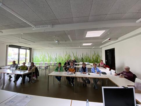 In einem gro&szlig;en Raum sitzen insgesamt 15 Menschen. Das Foto ist im Weitwinkel-Modus aufgenommen, damit alle Teilnehmende, die im Raum verteilt sitzen, abgebildet sind.