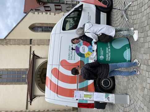 Theresa steht am Infostand vor dem Inbomobil und ber&auml;t zwei Seniorinnen. Im Hintergrund ist ein Dom zu sehen.
