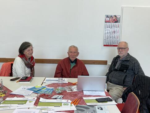 Die drei Teilnehmenden sitzen an einem mit Brosch&uuml;ren und Handouts gedeckten Tisch und l&auml;cheln in die Kamera.