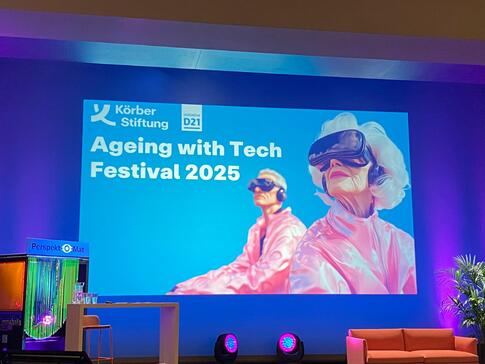 Eine B&uuml;hne im Theatersaal des K&ouml;rberHauses. Eine Pr&auml;sentation zeigt eine Folie mit der &Uuml;berschrift "Ageing with Tech Festival 2025". Rechts ist eine KI-generiertes Bild mit einer &auml;lteren Dama und einer j&uuml;ngeren Person, die eine VR-Brille tragen.