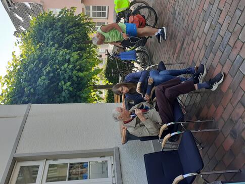 Auf dem Bild im Hochkantformat sitzt Petra mit einem &auml;lteren Herr auf zwei St&uuml;hlen. Sie erkl&auml;rt ihm etwas auf dem Smartphone. Rechts im Hintergrund steht ein sommerlich gekleideter Mann an einem Fahrrad.