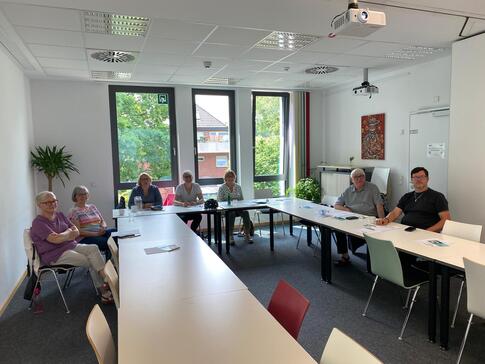 In einem Seminarraum sitzen an Tischen in U-Form f&uuml;nf Frauen und zwei M&auml;nner. Alle blicken nach vorne in die Kamera. Die alle Teilnehmenden der Veranstaltung lie&szlig;en sich fotografieren.