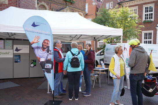 Vor einem gro&szlig;en Pavillon steht Petra mit einem Ehepaar. Links davon steht eine Beachflag der Volkshochschule Meppen und rechts im Bild unterh&auml;lt sich eine Frau mit einem Mann. Der Mann tr&auml;gt einen Fahrradhelm.