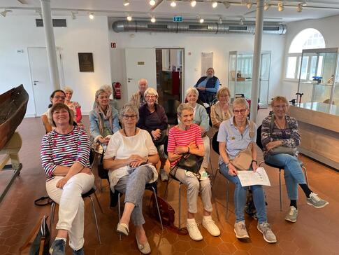 In einem Museumraum sitzen knapp f&uuml;nfzehn Senior:innen in drei Stuhlreihen und schauen nach vorne in die Kamera.