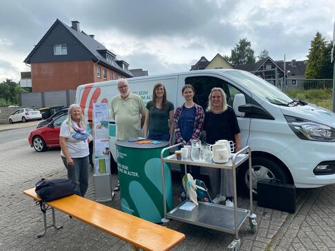 Am Info-Mobil steht Petra (Mitte) mit Anna (2. v. r.) und Maren Korban von der AWO (1. v. r.). Links von Petra steht ein AWO-Vorstandsmitglied und Annas Stammg&auml;stin. Vor den f&uuml;nf Personen steht der Info-Stand mit einer Bierbank und einem Wagen, auf dem Kaffee und Wasser steht.
