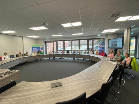 Im modernen Rathaussaal sitzen in einer gro&szlig;en ovalen Form mit Tischen ungef&auml;hr 17 Personen. Sie sind schwer zu erkennen, weil das Bild im Weitwinkelformat vor allem den Tisch vorne zeigt.