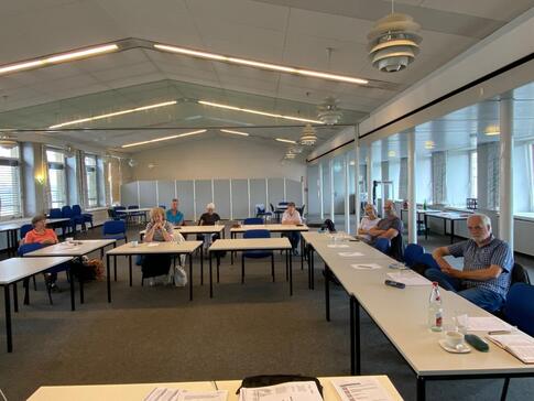 In einem sehr gro&szlig;en Veranstaltungsraum sitzen acht Personen an Tischen in U-Form. Zwei Personen wollten nicht fotografiert werden.