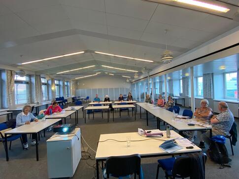 In einem sehr gro&szlig;en Raum sitzen ca. 13 Personen an Tischen, die in einer U-Form aufgestellt sind. Im Vordergrund steht ein Tisch mit einem Laptop und ein kleiner Schrank mit einem Beamer.