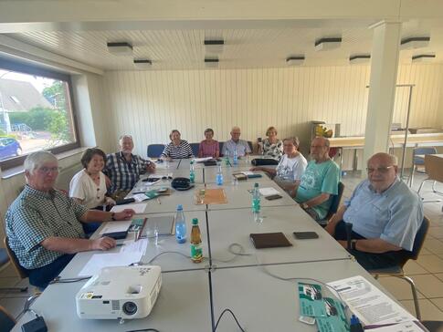 Elf Senior:innen sitzen im D&ouml;rphus an einem gro&szlig;en, rechteckigen Tisch. Sie blicken nach vorne in die Kamera. Auf dem Tisch liegen Unterlagen, Wasserflaschen und ein Beamer.