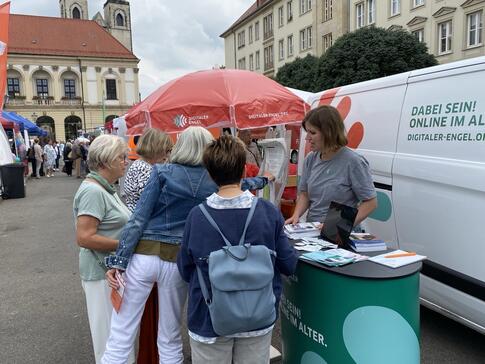 Mobilreferentin Theresa erkl&auml;rt 4 Seniorinnen etwas am Infostand auf dem Marktplatz. Rechts neben ihr ist das Infomobil.