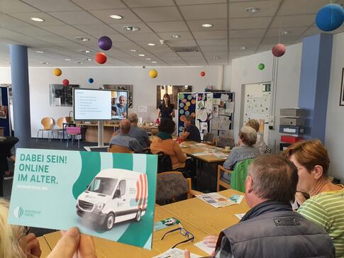 Links im Vordergrund ist ein Flyer vom Digitalen Engel in der Luft gehalten. Rechts davon und im Hintergrund sitzen &auml;ltere Personen an Tischen. Ganz weit im Hintergrund steht Petra neben einer digitalen Tafel.