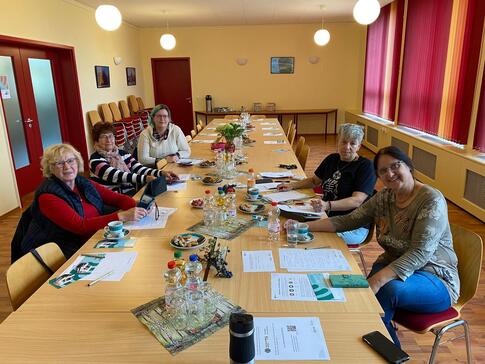 F&uuml;nf Frauen sitzen an einem Tisch gegen&uuml;ber und schauen in die Kamera nach vorne. Auf dem Tisch liegen Materialien und Herbstdeko. 