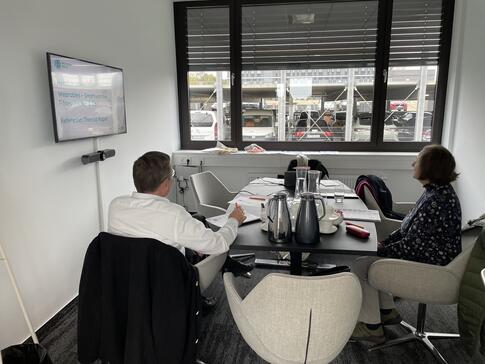 Zwei Teilnehmende sitzen in einem kleineren Besprechungsraum an einem Tisch. Sie sind von hinten oder von der Seite zu sehen. Links im Raum ist der Monitor mit der ersten Folie der Pr&auml;sentation. Im Hintergrund ist ein Fenster, durch das Autos zu sehen sind.