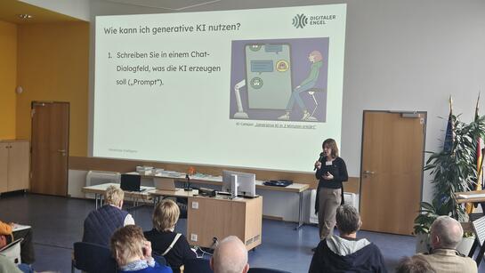 Theresa steht rechts neben einer gro&szlig;en Leinwand, auf der eine Folie zu generativer KI zu sehen ist. Theresa spricht mit Mikrofon in der Hand. Teilnehmende in den vordersten Reihen sind von hinten zu sehen.