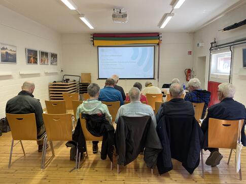 Die 10 Teilnehmenden und der Kooperationspartner sitzen in drei Stuhlreihen hintereinander, sind von hinten zu sehen und schauen auf die Leinwand mit der Startfolie der Pr&auml;sentation.