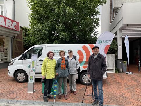 4 Besucher:innen stehen in einer Einkaufsstra&szlig;e vor dem Infomobil am Infostand und l&auml;cheln in die Kamera. Der Himmel ist dunkel aufgrund bevorstehenden Regens. 
