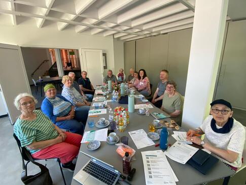 An einem l&auml;nglichen Tisch sitzen zirka 10 Personen und blicken nach vorne in die Kamera. Auf dem Tisch liegen Unterlagen, Getr&auml;nke und Geb&auml;ck.