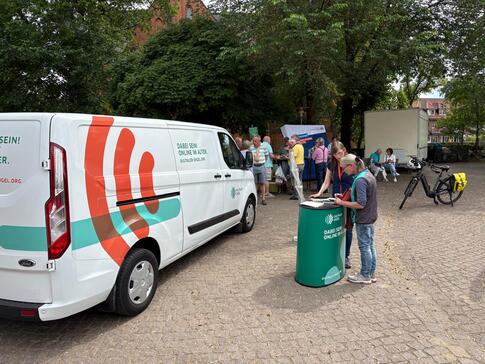 Links im Bild steht das info-Mobil. In der Mitte am Info-Stand Petra mit einer Seniorin. Im Hintergrund stehen weitere Personen und unterhalten sich.