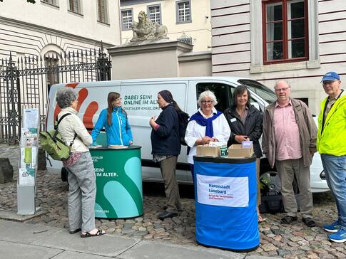 Auf der rechte Seite ist ein Messetresen vom Seniorenbeirat L&uuml;neburg mit vier Senior:innen, die in die Kamera posieren. Auf der linke Seite ist Referentin Petra mit zwei Damen im Gespr&auml;ch. Im Hintergrund steht das Info-Mobil.
