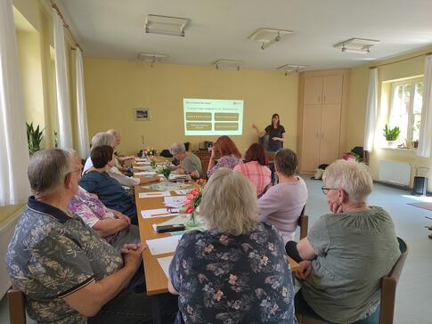 An einem langen Tisch sitzen (von hinten fotografiert) Menschen. Sie blicken nach vorne auf Petra, die neben einer Pr&auml;sentation steht.
