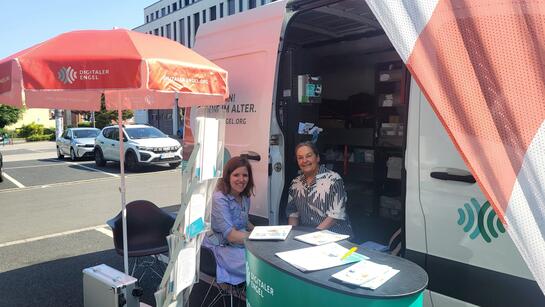 Theresa und eine &auml;ltere Besucherin sitzen am Infostand auf einem Supermarktparkplatz.