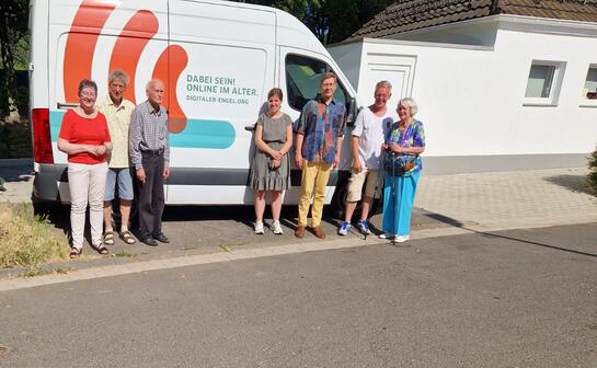 Theresa, Ehrenamtliche und Besucher:innen stehen bei Sonnenschein vor dem Infomobil links neben einem wei&szlig;en Geb&auml;ude.