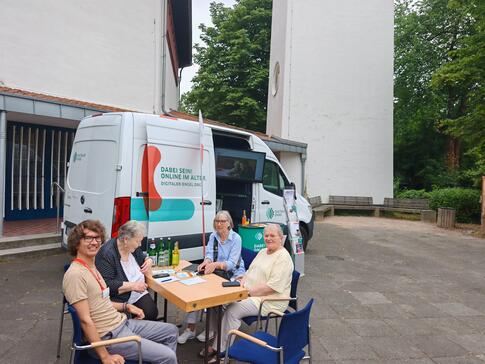 Referent Johannes sitzt mit drei Besucherinnen an einem Tisch vor dem Infomobil.