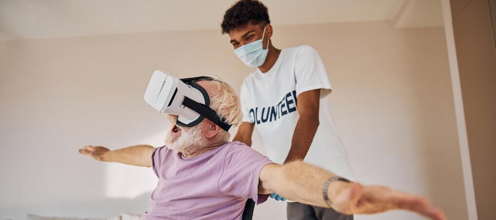 &Auml;lterer Herr im Rollstuhl tr&auml;gt eine VR Brille und wird von einem jungen l&auml;chelnden Mann geschoben.